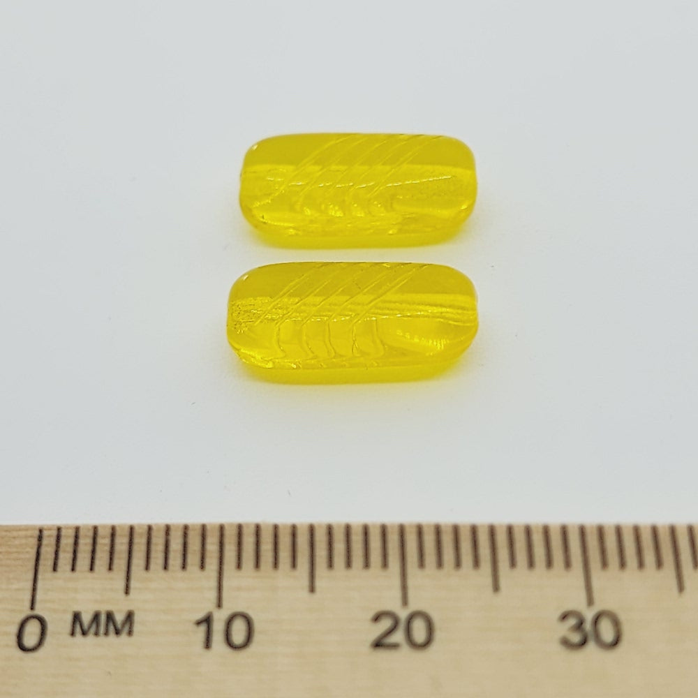 close up of 2 vibrant yellow clear beads shaped like a pillow with a ruler to show scale on a white background