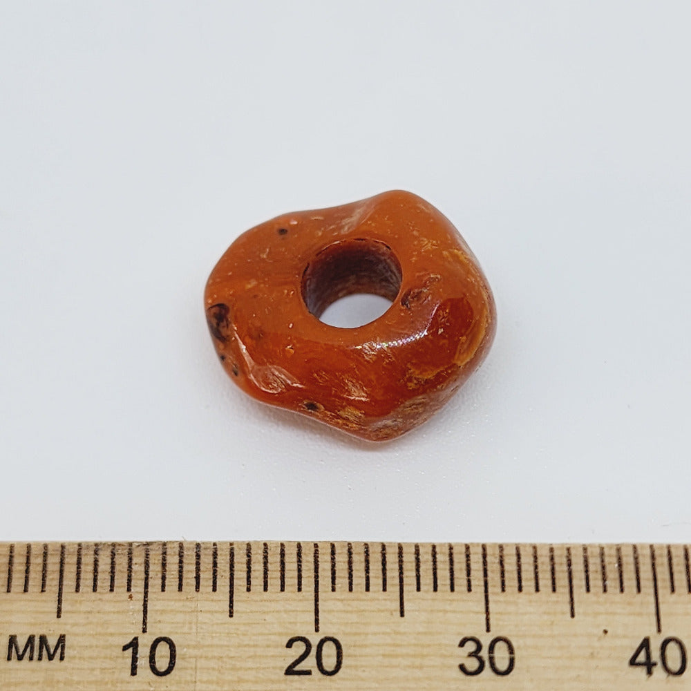 Orange or brown bead with a big threading hole next to a wooden ruler on a white background