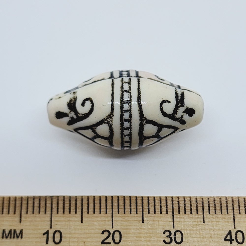 A white and black torpedo-shaped lucite bead with geometric Art Deco style etchings, placed next to a ruler for scale.