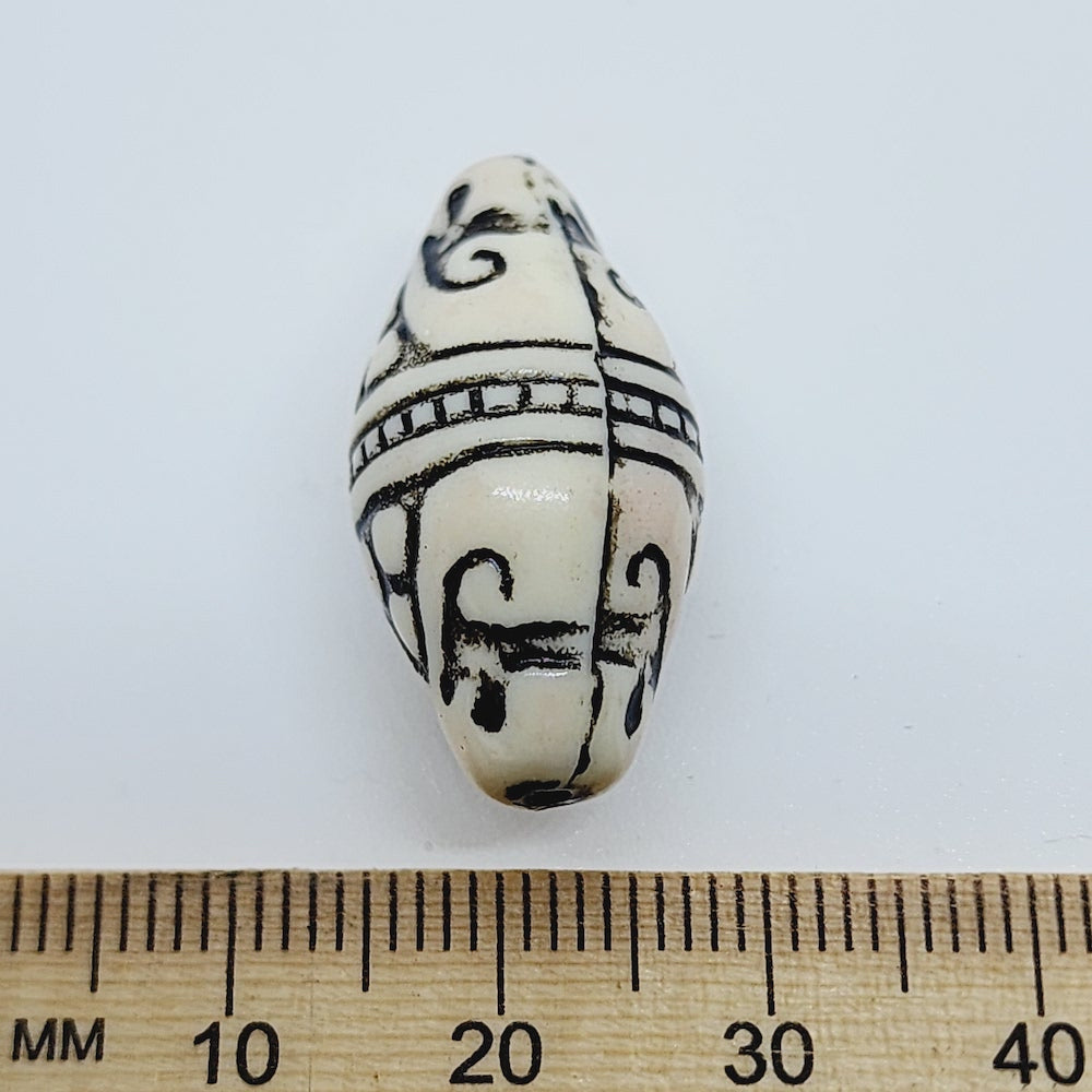 A white and black bicone-shaped lucite bead with geometric Art Deco style etchings, placed next to a ruler for scale.