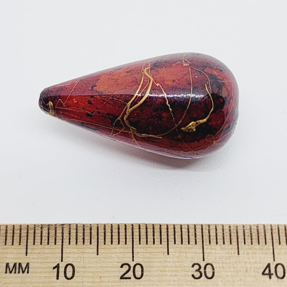 A teardrop-shaped vintage Lucite bead with a rust bubble glaze look and gold drizzle patterns, next to a ruler for scale.