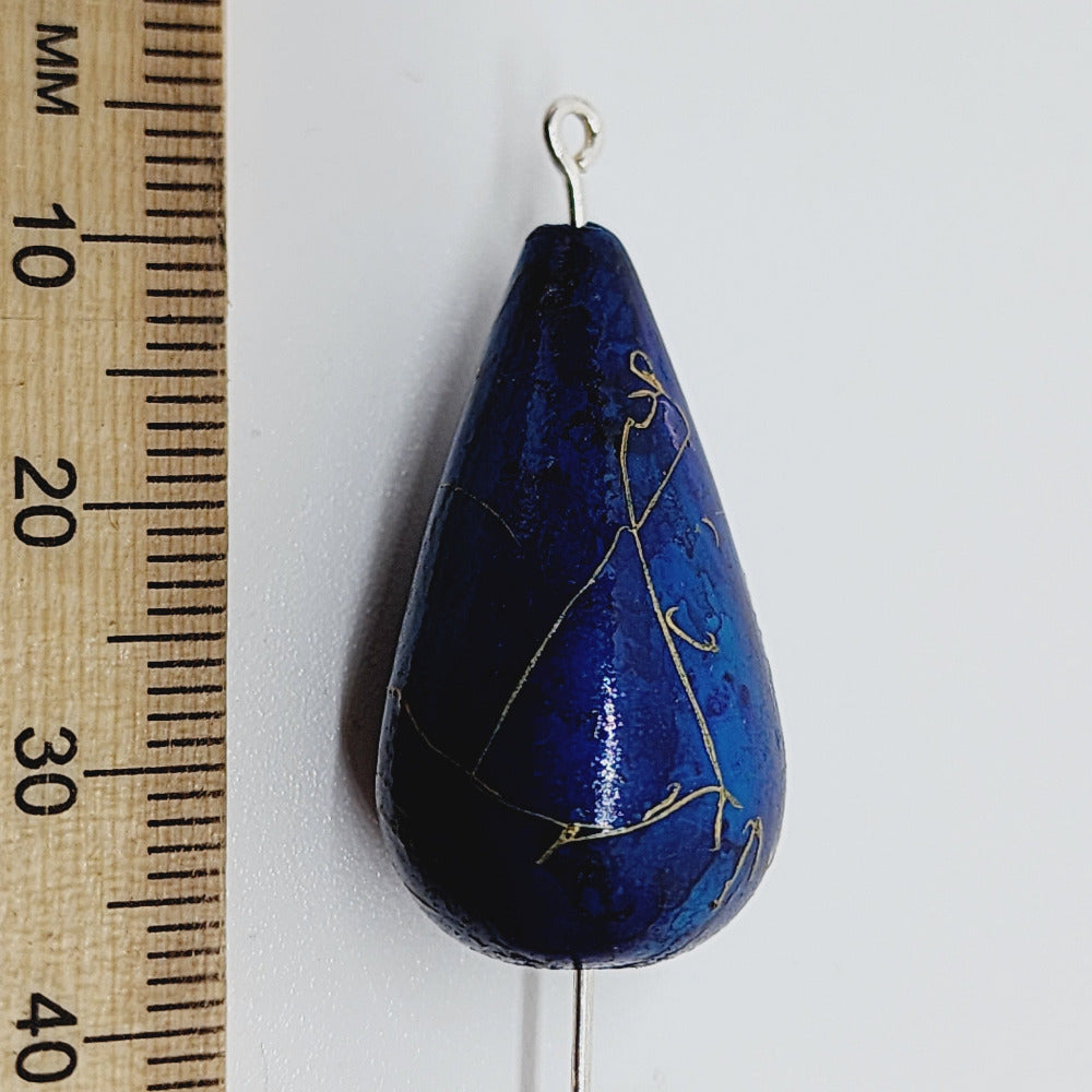 A teardrop-shaped vintage Lucite bead in blue with a gold drizzle pattern, next to a ruler for scale.