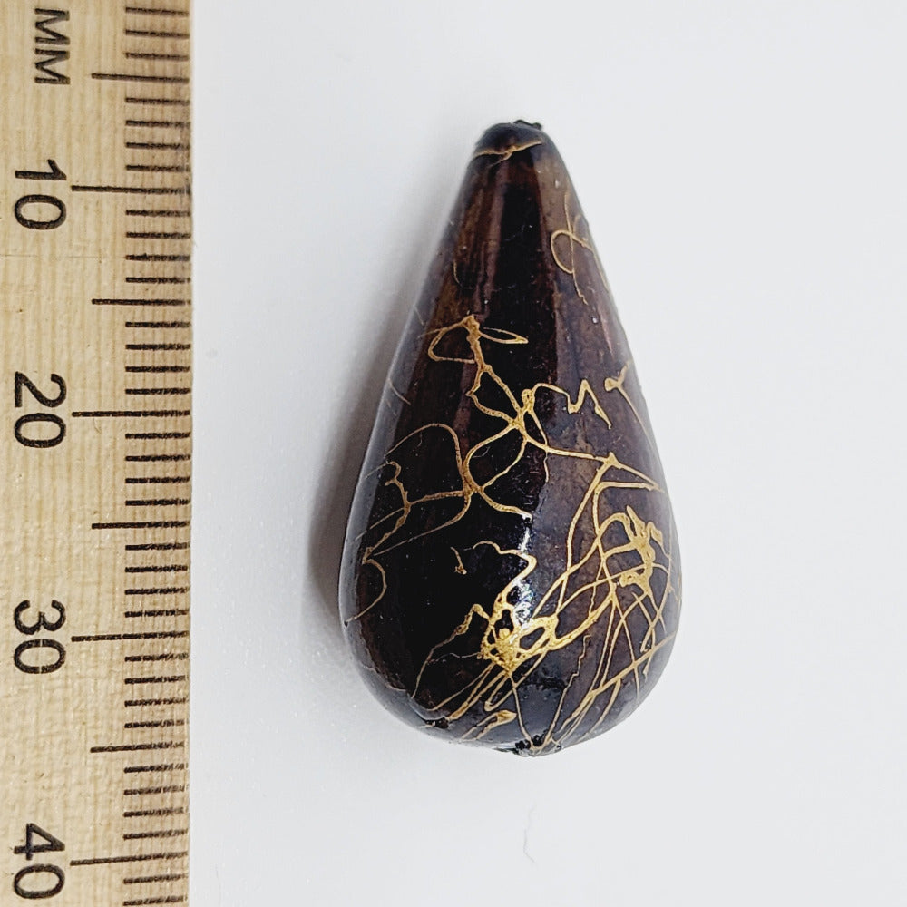 A teardrop-shaped vintage Lucite bead in brown with a gold drizzle pattern, next to a ruler for scale.