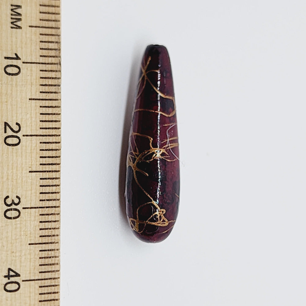 A teardrop-shaped vintage Lucite bead with a red bubble glaze look and gold drizzle patterns, next to a ruler for scale.