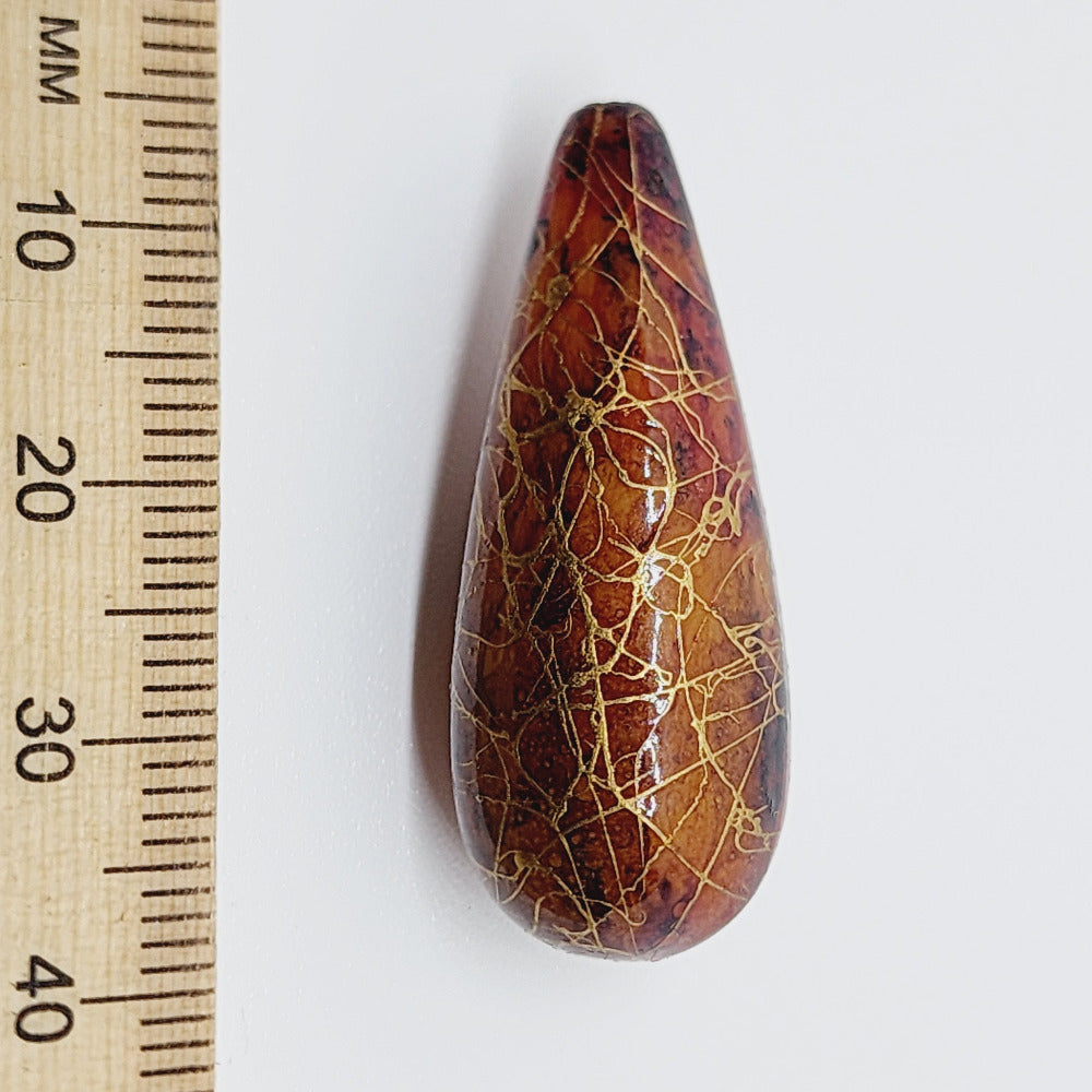 A teardrop-shaped vintage Lucite bead with a brown bubble glaze look and gold drizzle patterns, next to a ruler for scale.