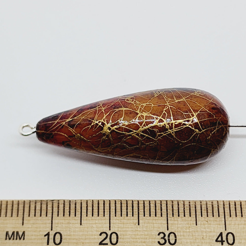 A teardrop-shaped vintage Lucite bead with a brown bubble glaze look and gold drizzle patterns, next to a ruler for scale.