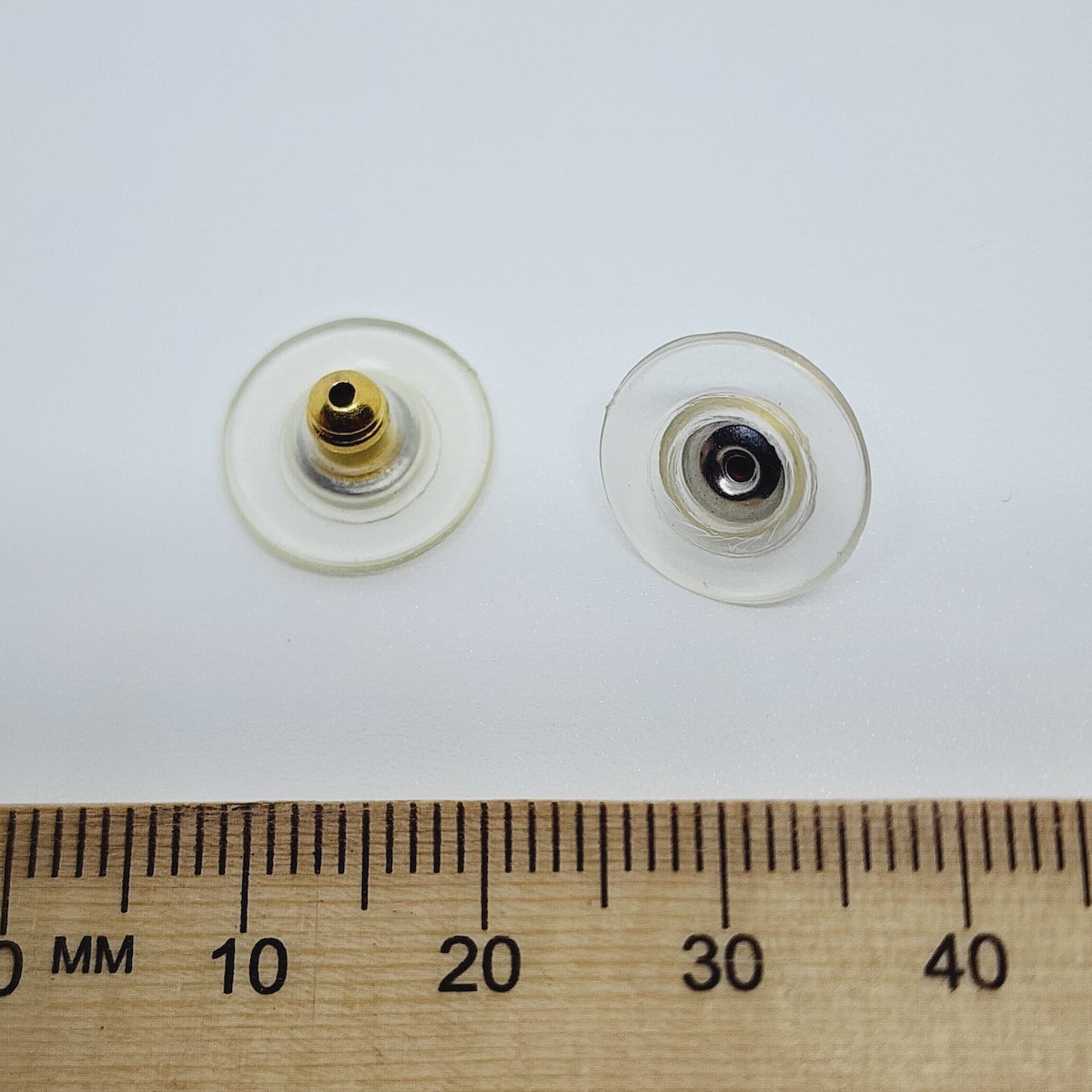 Two gold-colored plastic disc ear stud backs with a central bullet fitting, displayed next to a wooden ruler for scale.