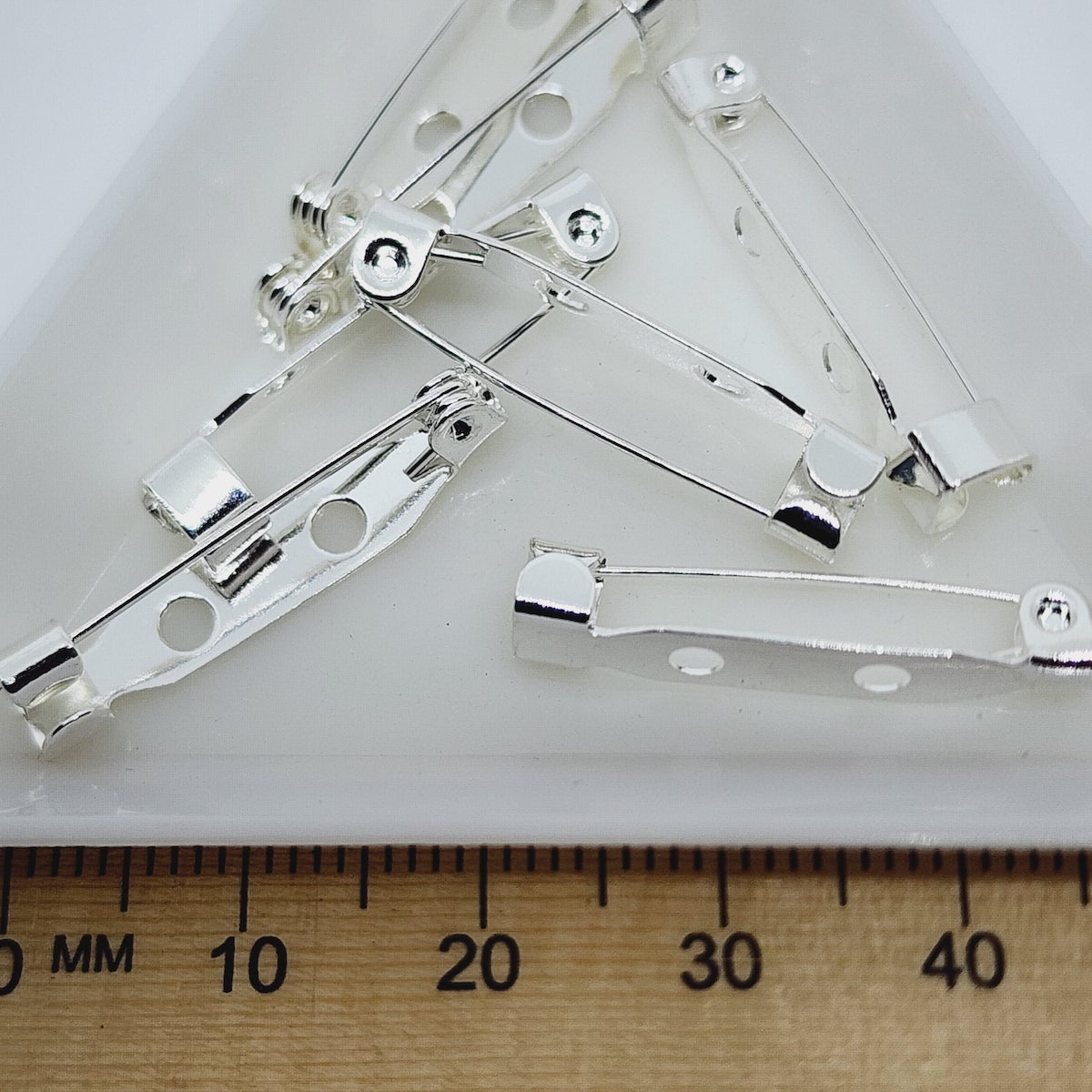 Silver brooch backs with two holes for attaching, displayed on a white surface next to a wooden ruler for scale.