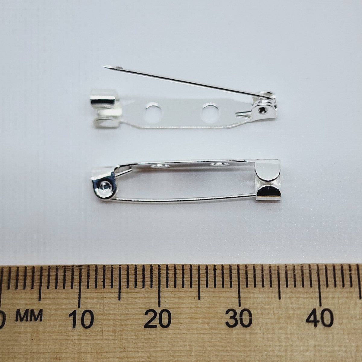 2 gold brooch back fittings on a white surface with a wood ruler to show size
