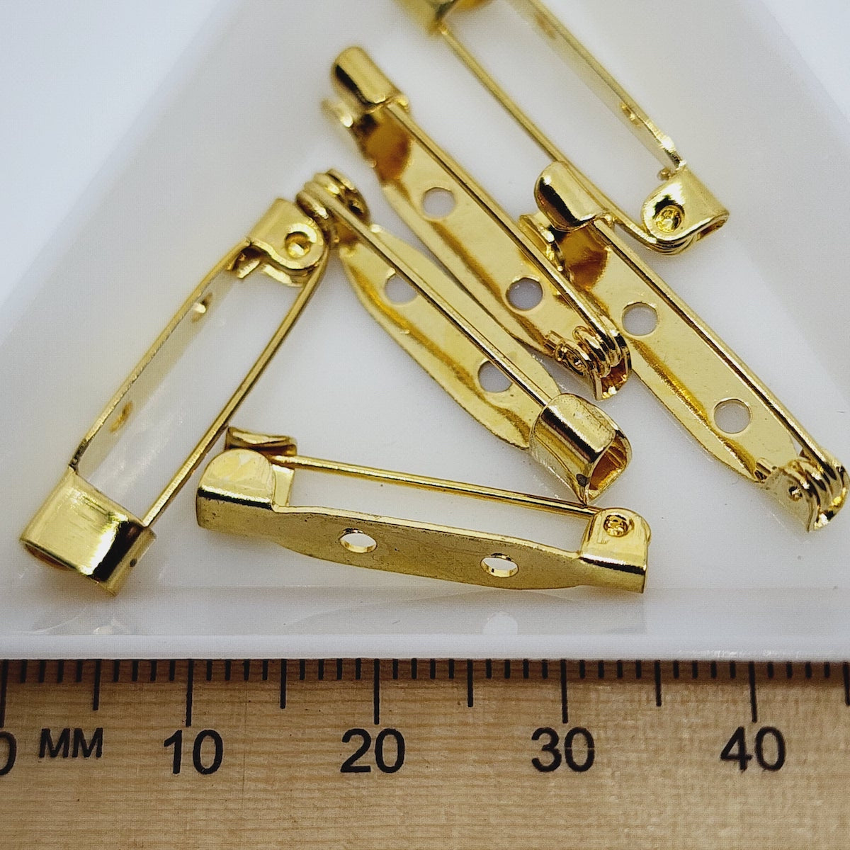 A set of gold-colored brooch backs laid out on a white surface, with a ruler at the bottom showing the scale in millimeters.