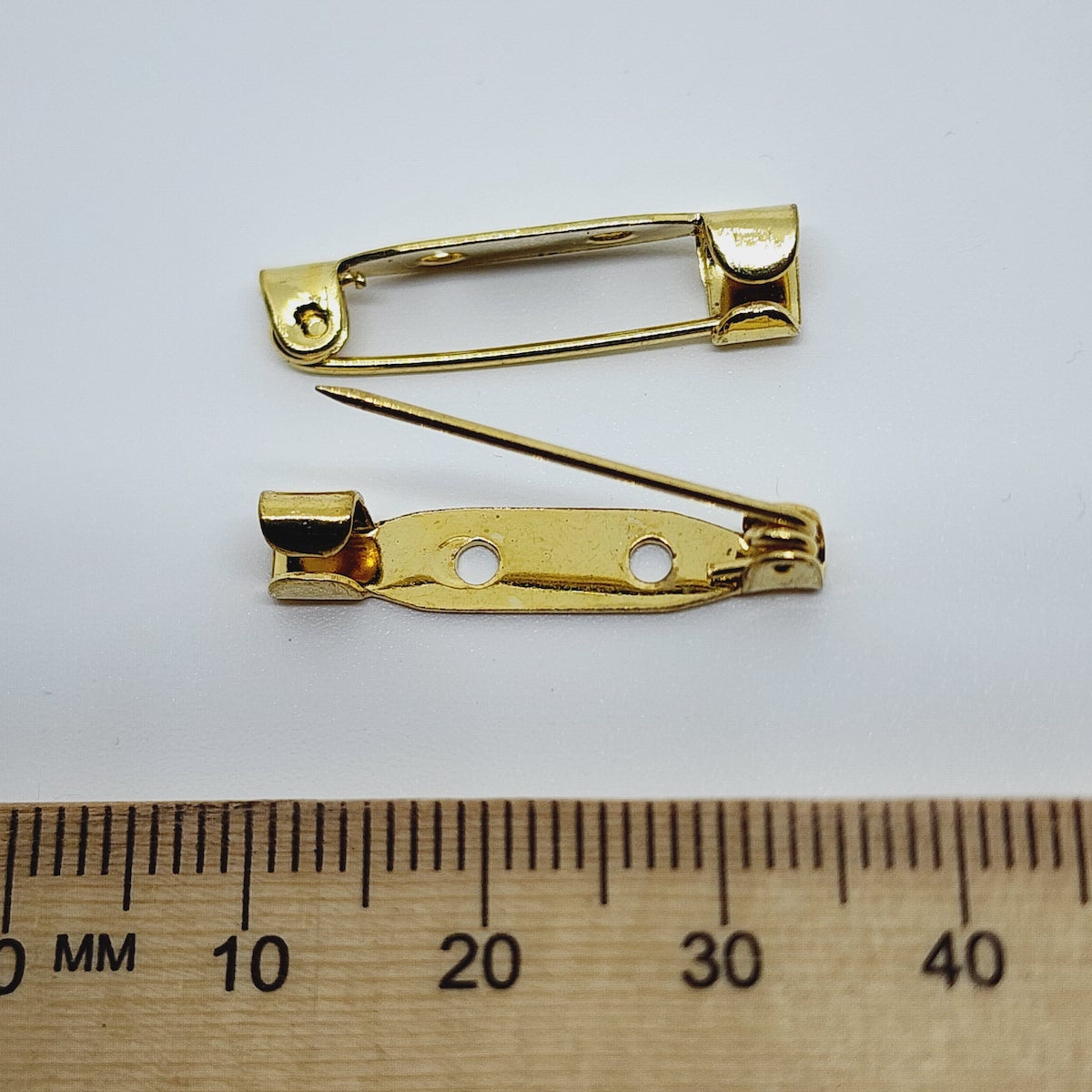 2 gold brooch back fittings on a white surface with a wood ruler to show size