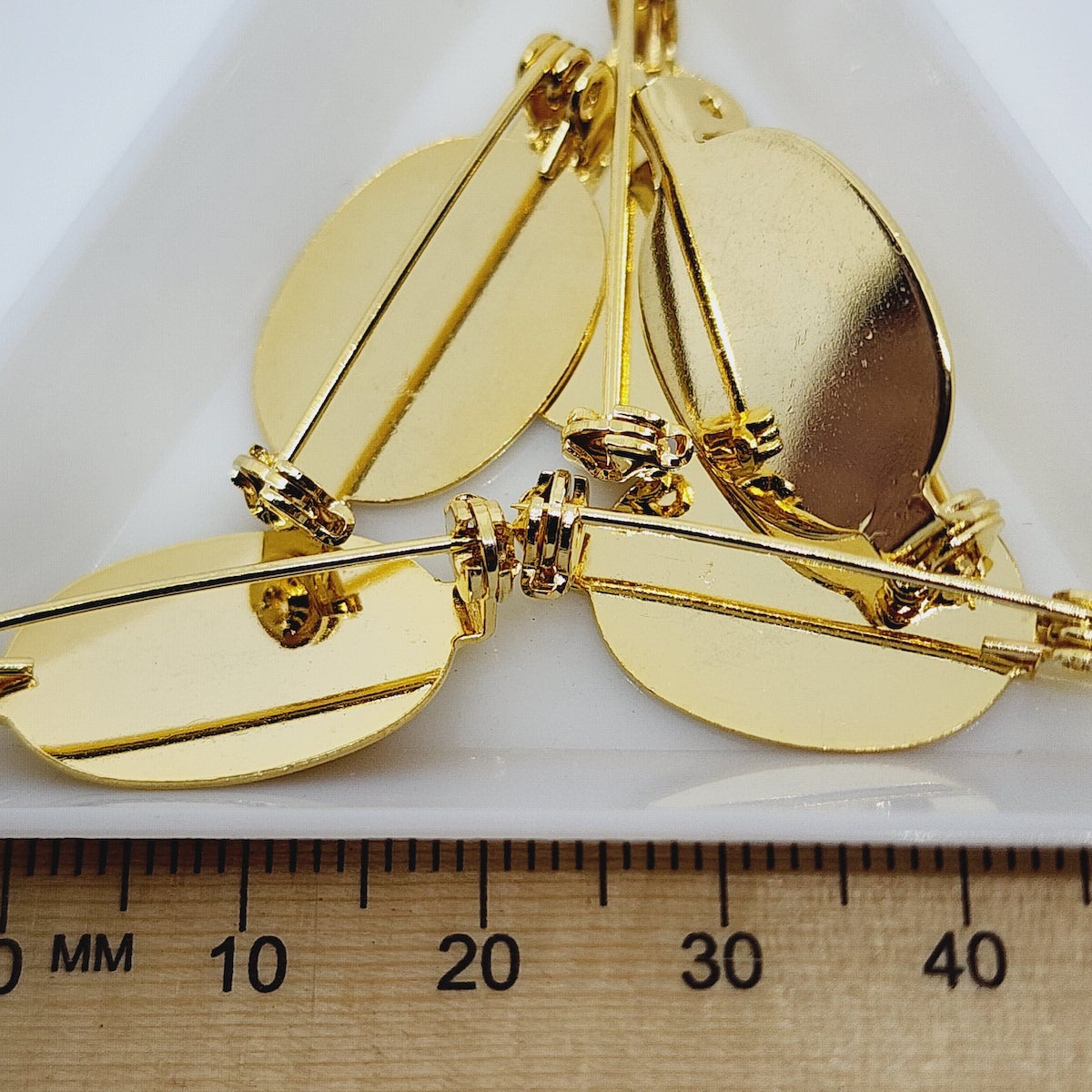 A set of gold-colored brooch settings with flat pad surfaces, displayed alongside a ruler for scale.
