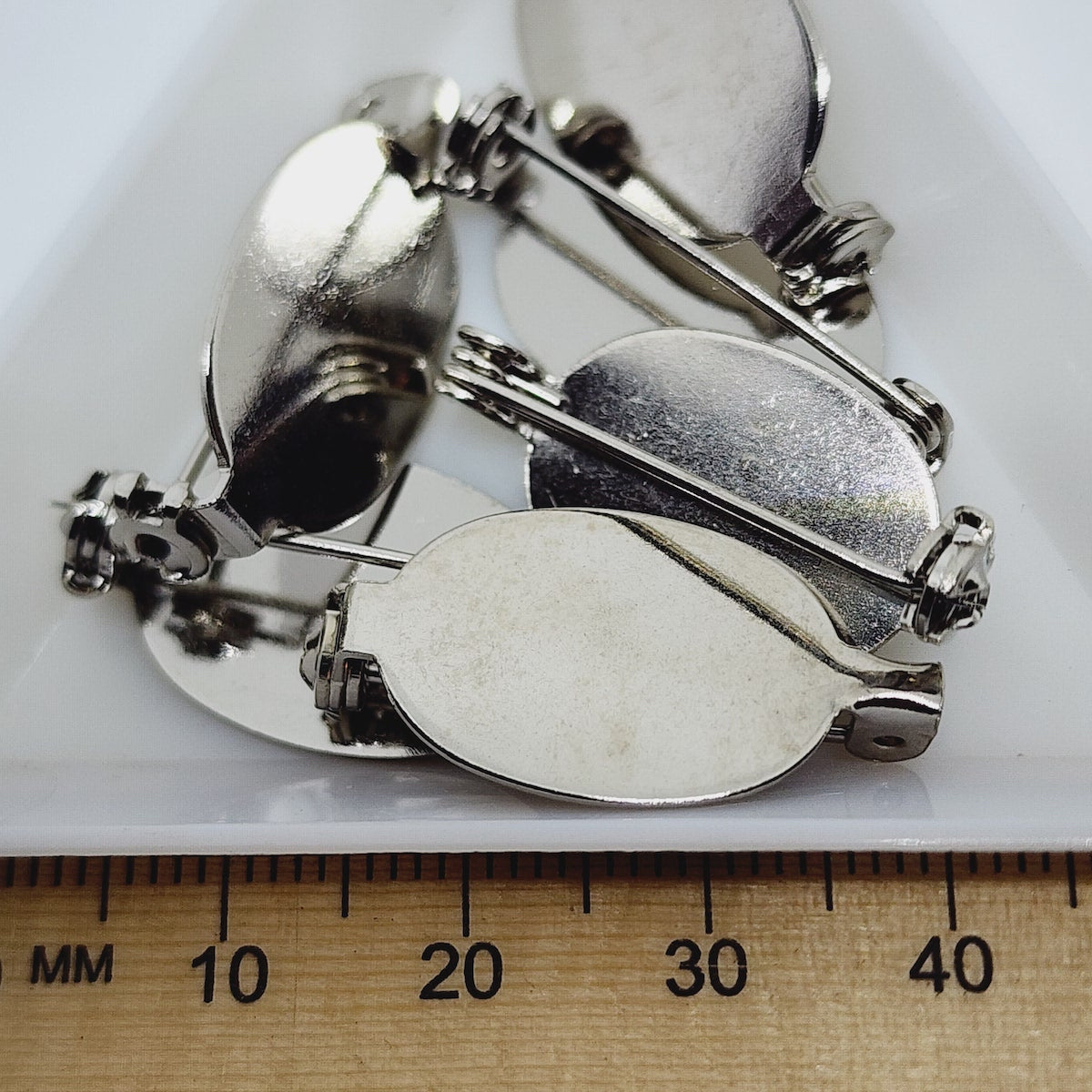 A set of silver brooch settings on a white surface, with a ruler showing the settings to be approximately 20x14mm in size.