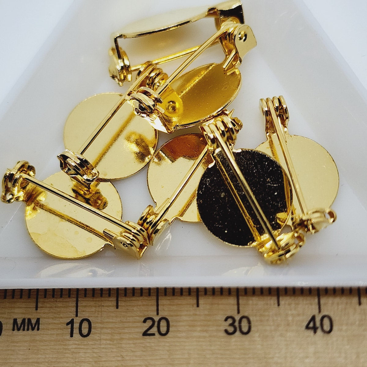 A collection of gold brooch settings displayed on a white surface, with a ruler at the bottom for scale.