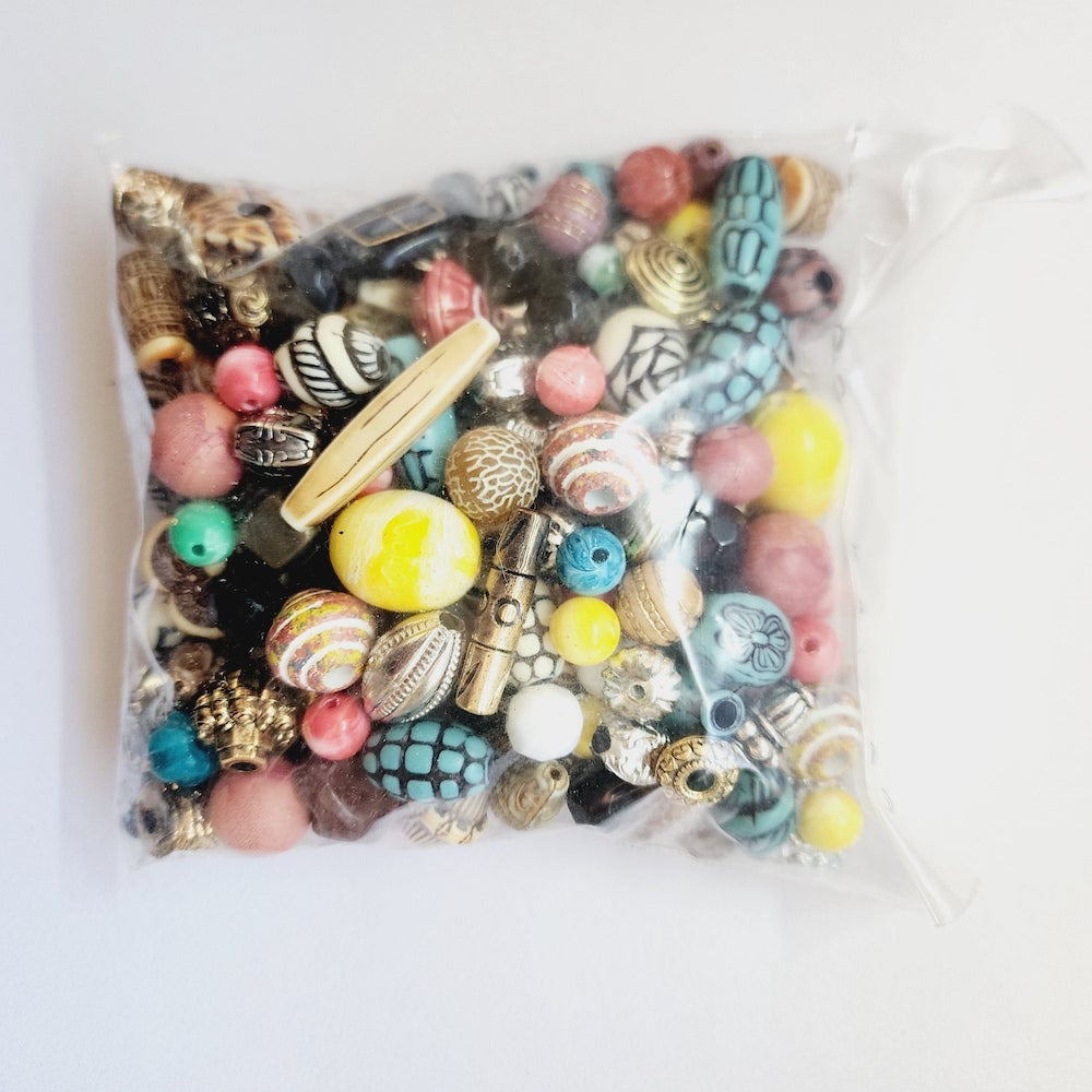 A clear plastic bag filled with assorted decorative beads and charms in many colors, including pink, yellow, turquoise, blue, black, white, and metallic gold, photographed on a plain white background.
