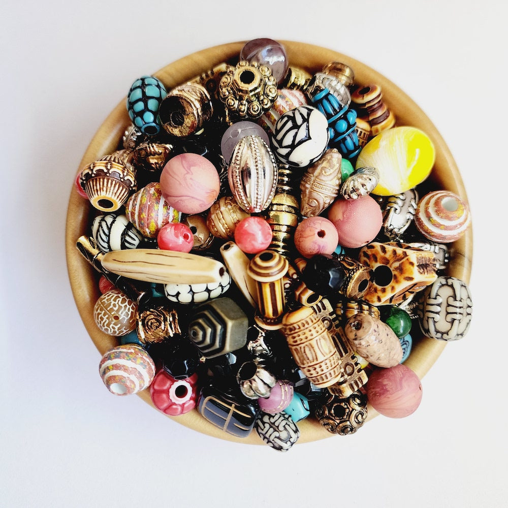 A top-down view of a wooden bowl filled with an assortment of colorful, patterned beads and decorative jewelry components on a plain white background.