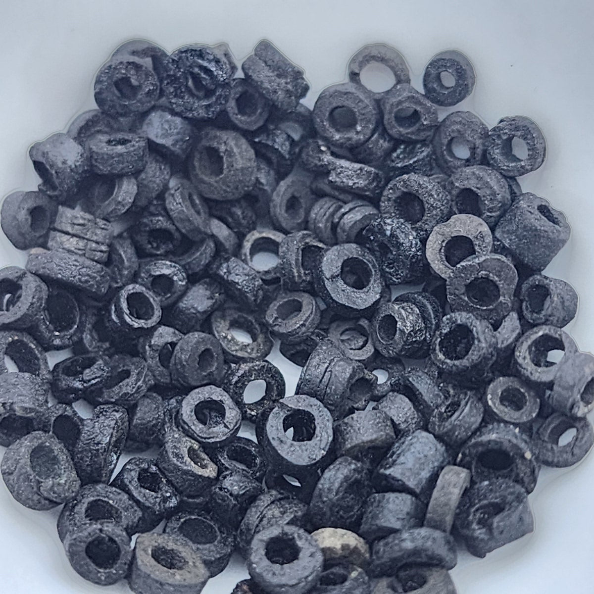 A close up photo of a collection of black vintage faience clay beads on a white background showing different shapes and sizes with a clear look at the threading holes and quality