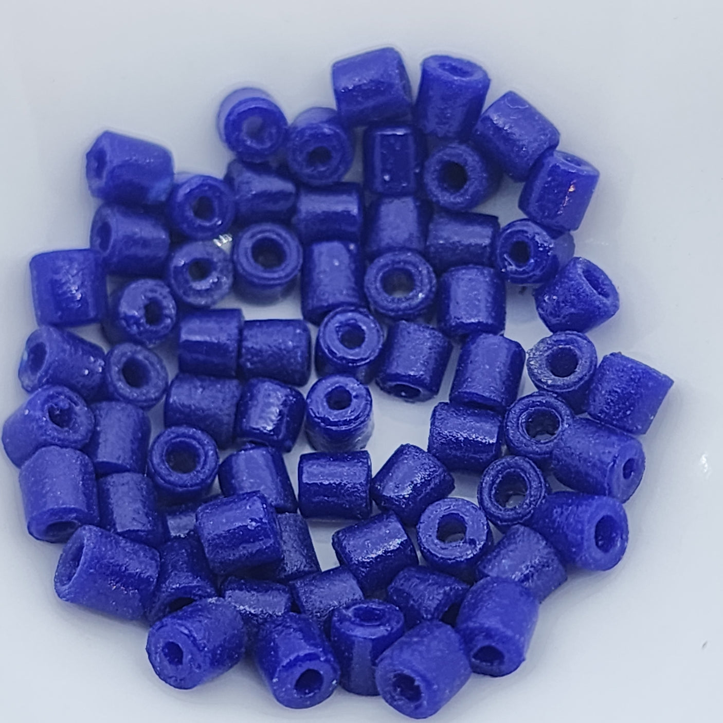 A collection of vintage Egyptian lapis lazuli blue cylindrical glass beads made from powdered glass with visible holes for stringing, on a light background showing visible holes for stringing