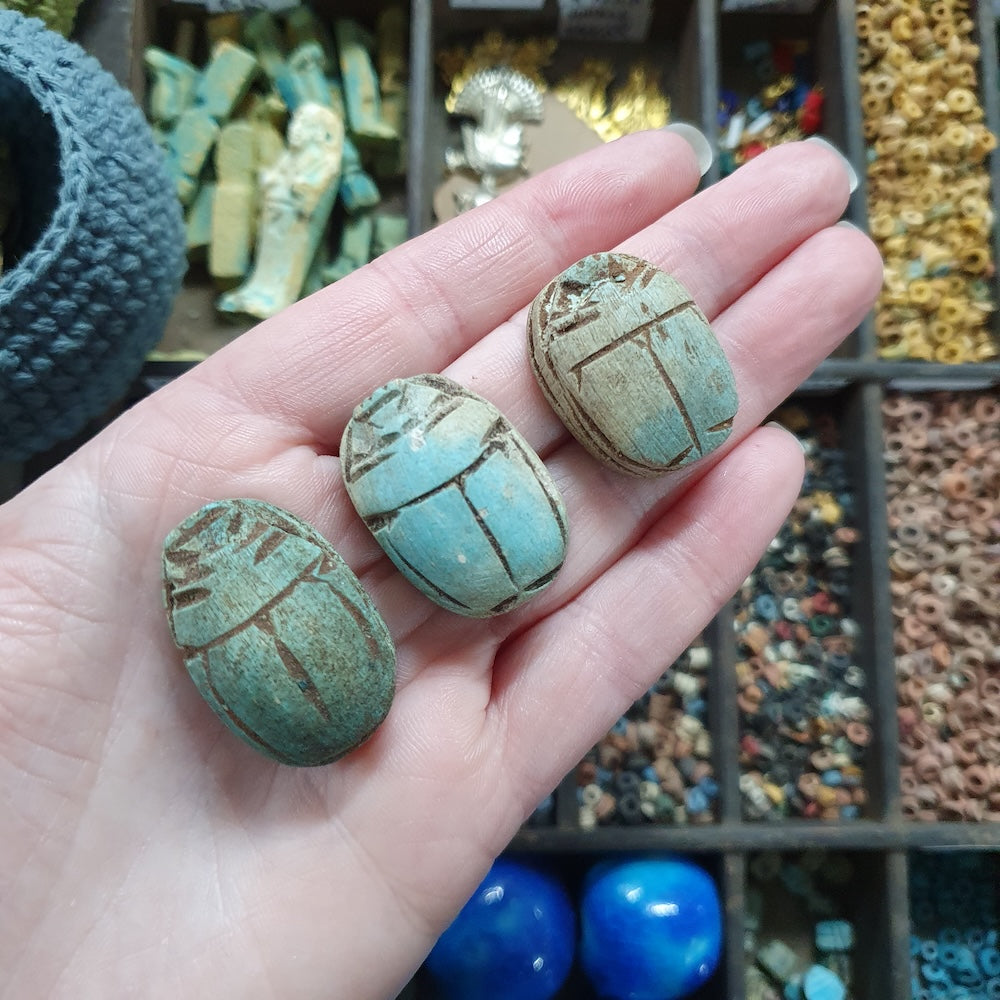 Three turquoise stone scarabs held in a hand with a display of similar items in the background.