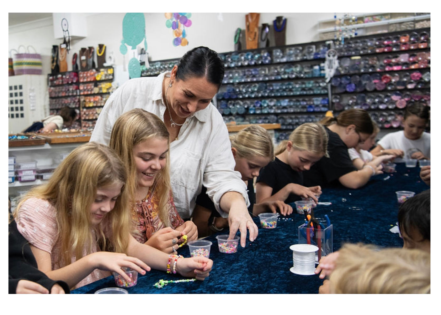 Table showing lots of children doing a beading workshop with a shop background with jars of beads and cord reels