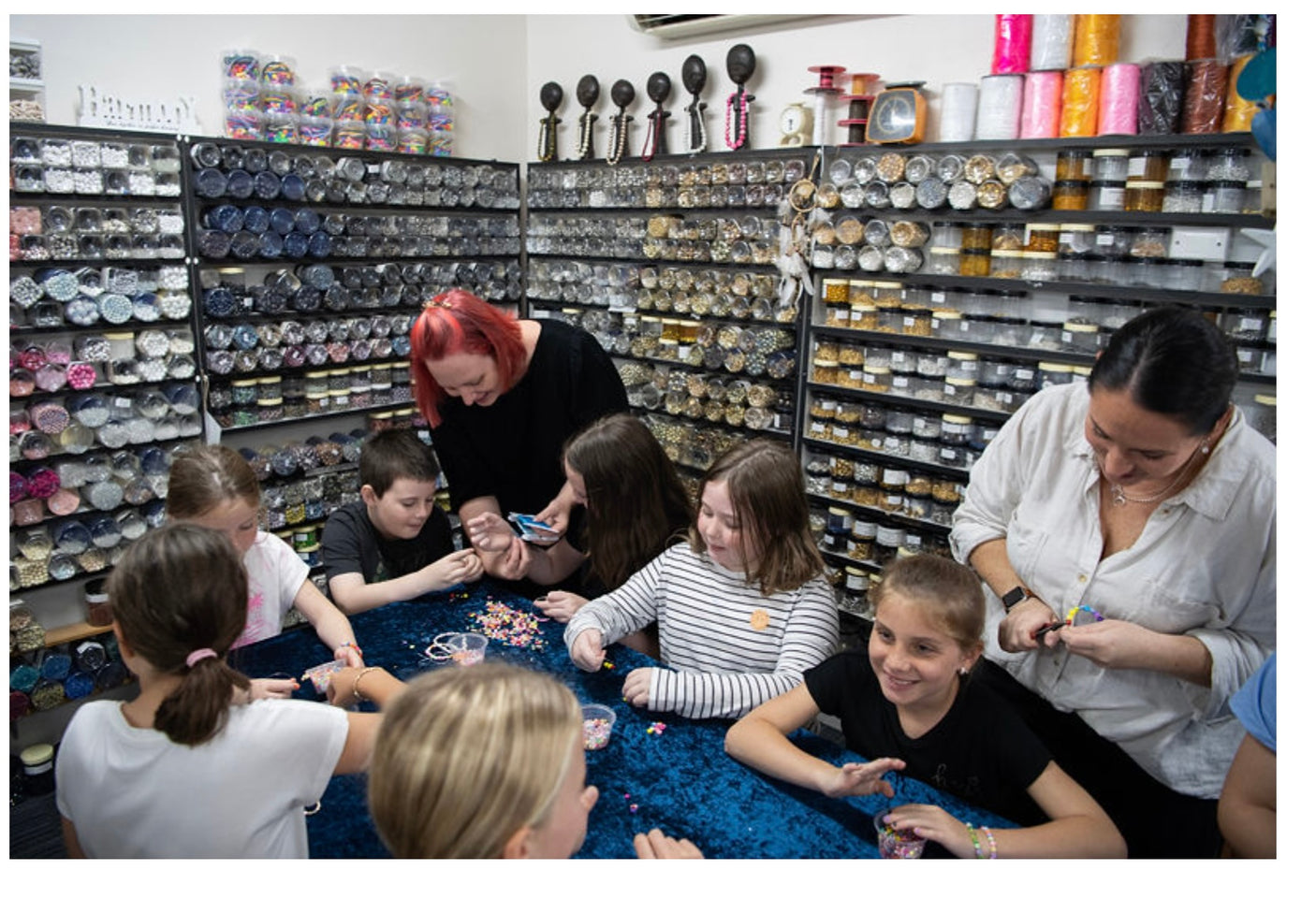 Table showing lots of children doing a beading workshop with a shop background with jars of beads and cord reels
