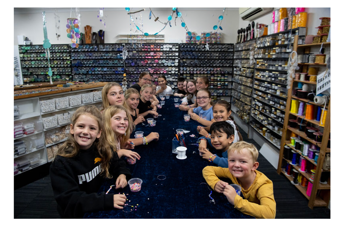 Table showing lots of children doing a beading workshop with a shop background with jars of beads and cord reels