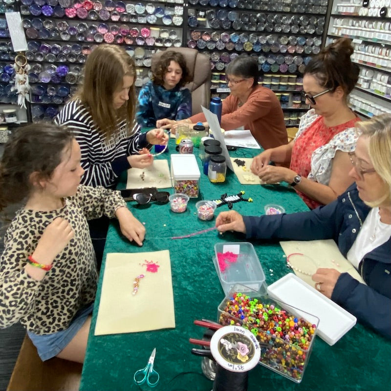 Table showing lots of children doing a beading workshop with a shop background with jars of beads and cord reels on the table with tools