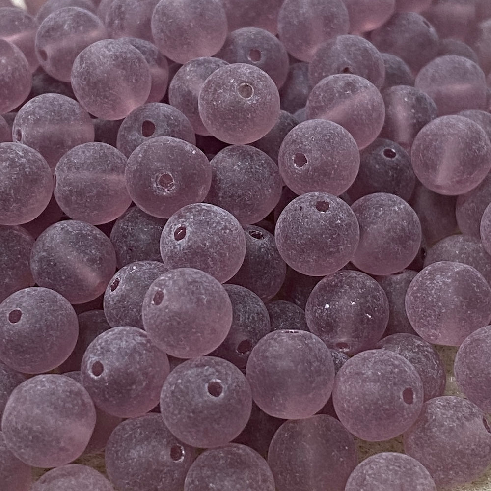 A close-up pile of small, round, frosted glass beads in a dusty lavender-purple color, each featuring a pre-drilled hole.