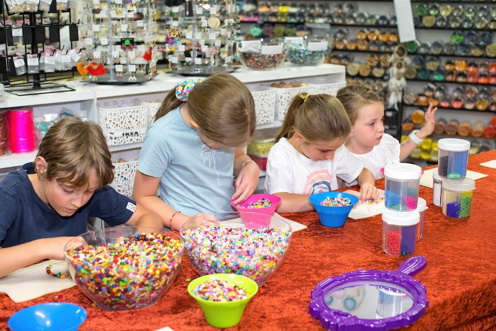 four children beading at a work table at Bead Shack using big bowls of mixed colourful beads with a mirror on the table and jars and bowls of beads in the background