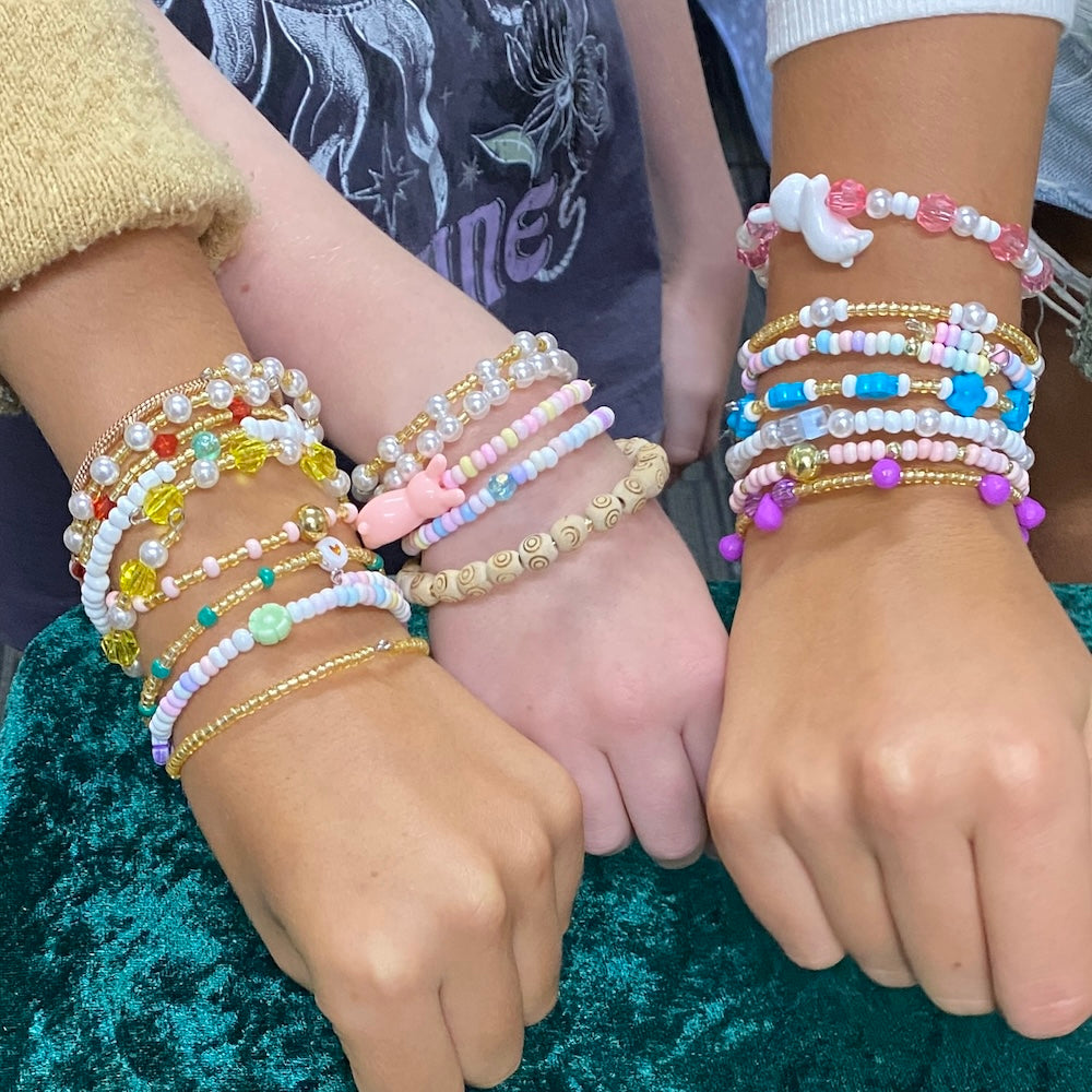 Three hands wearing colorful beaded bracelets on a textured green surface.