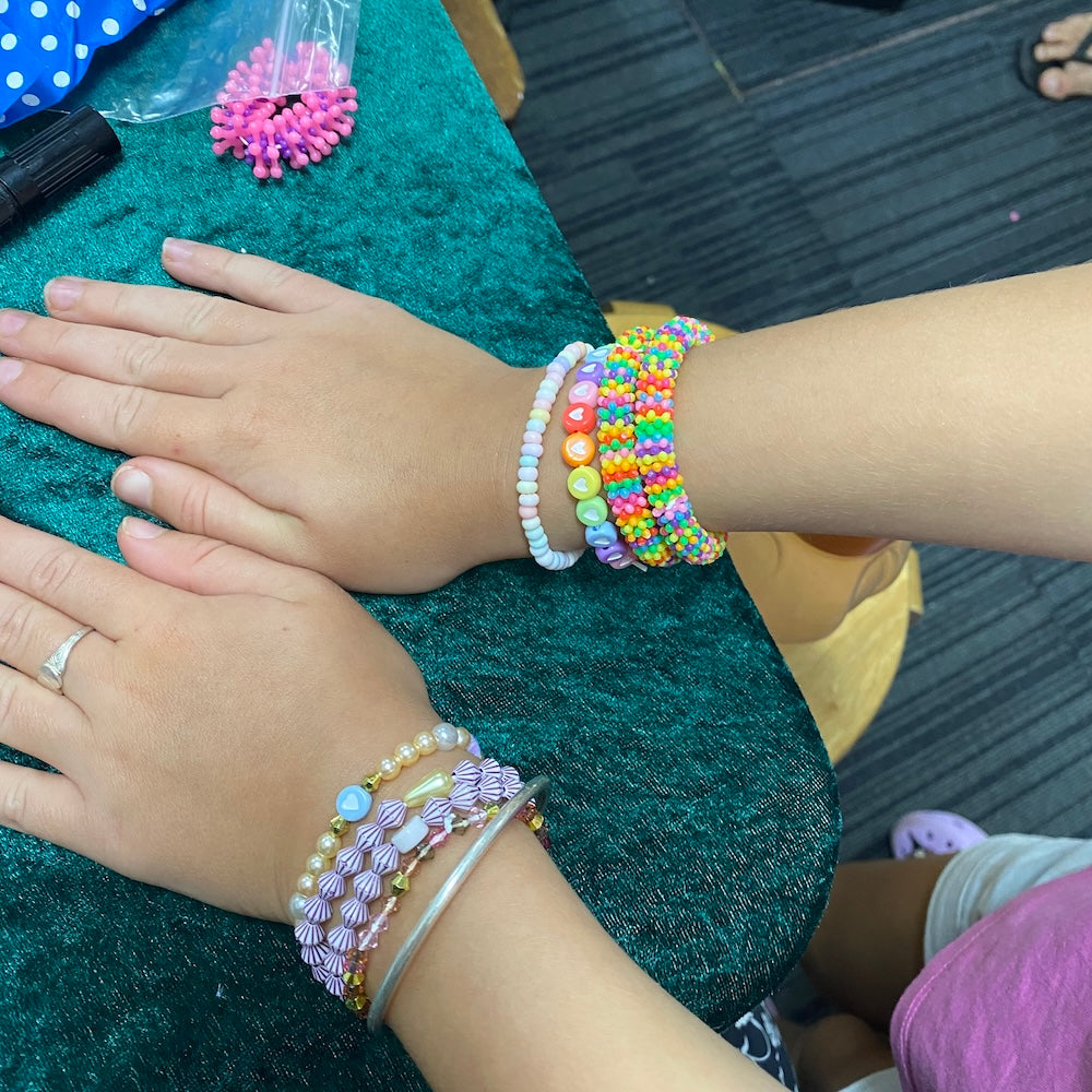 Two hands wearing colorful beaded bracelets on a green cushion.