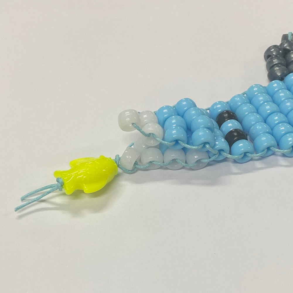shark toy beaded from a kit in blue and grey pony beads with white beads for teeth and a fish in the mouth on a grey textured background