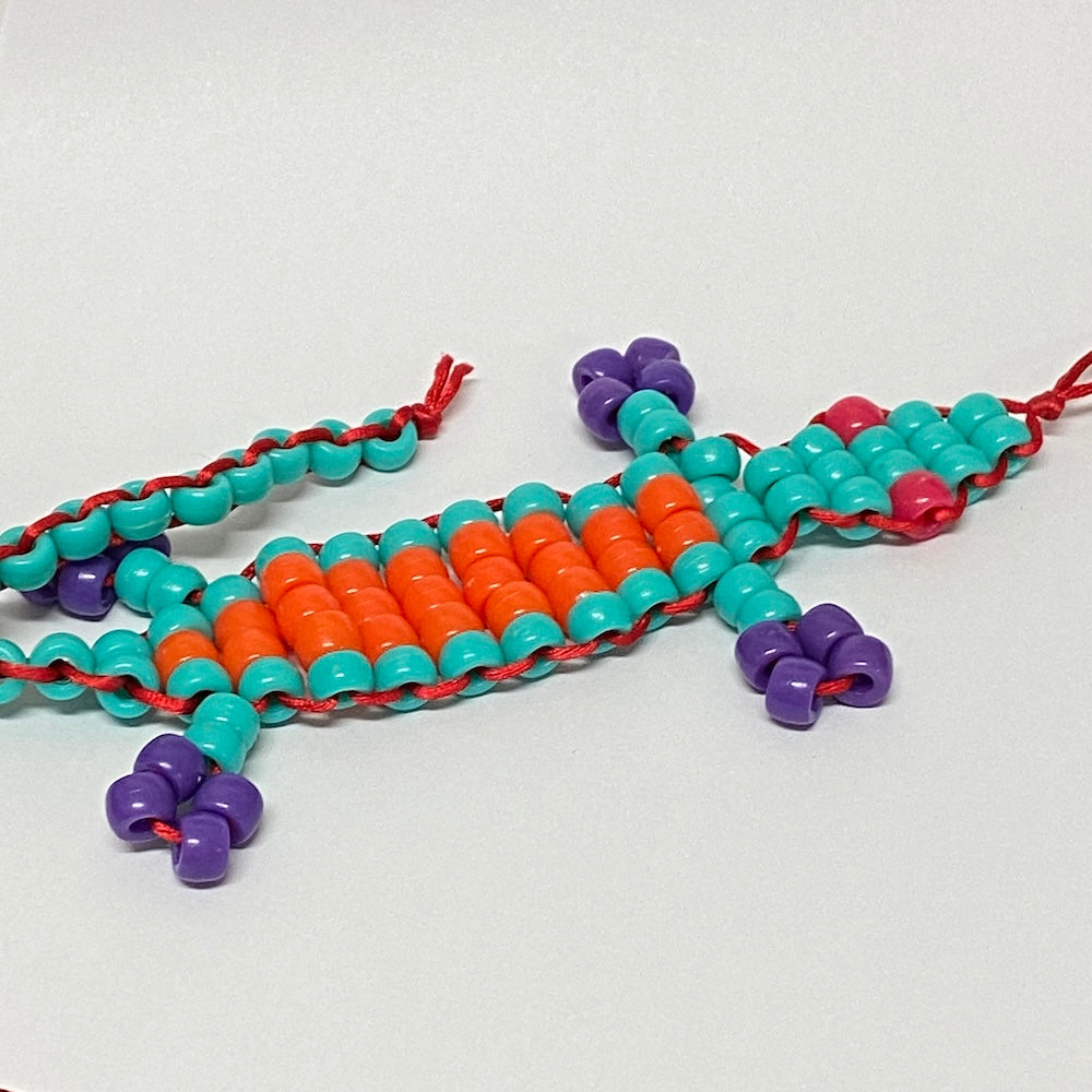 A handmade toy beaded gecko kit with orange, teal, and purple beads on a white background.