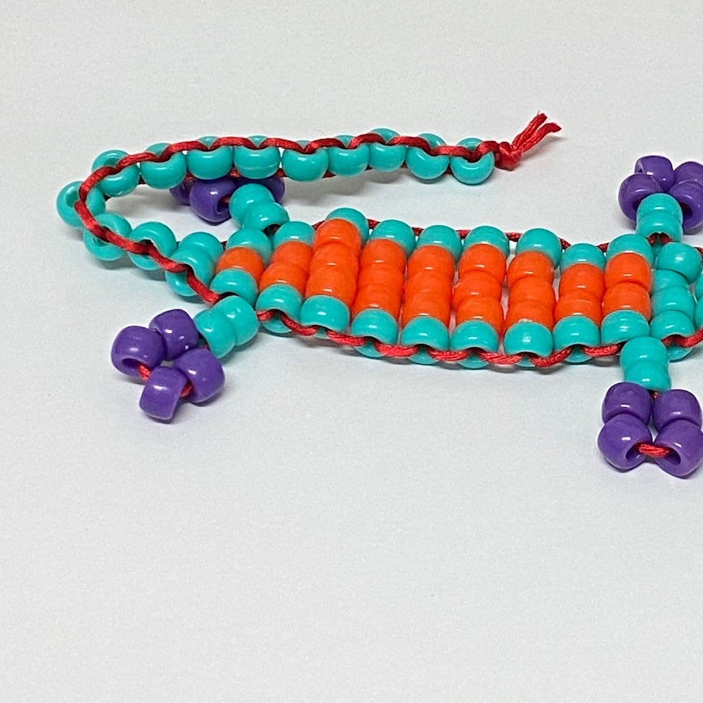 A handmade toy beaded gecko kit with orange, teal, and purple beads on a white background.