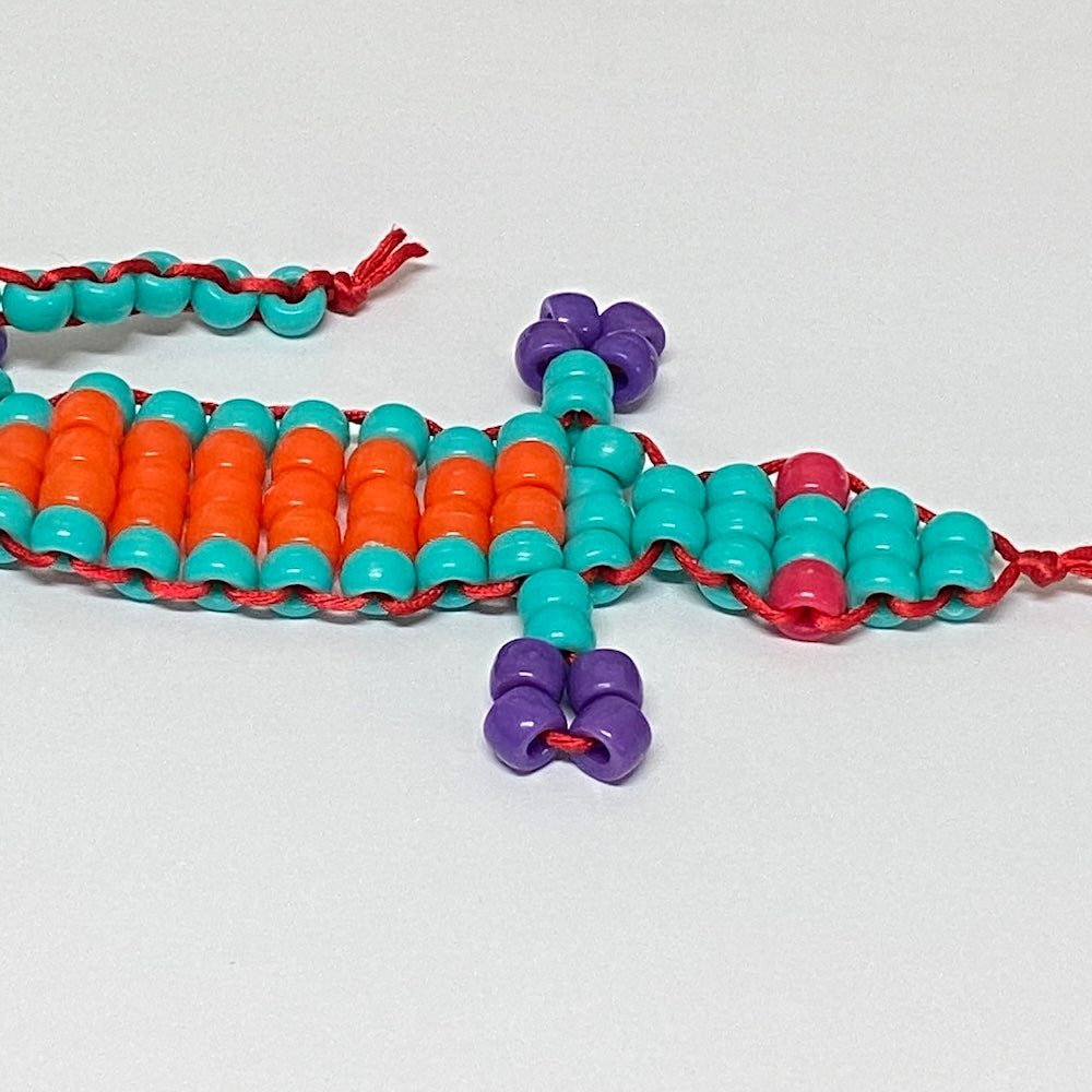 A handmade toy beaded gecko kit with orange, teal, and purple beads on a white background.