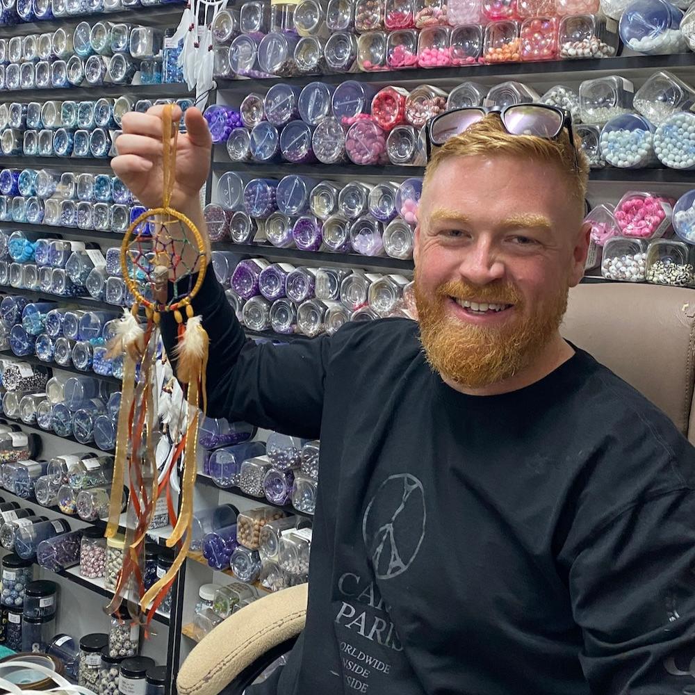 Finished dreamcatcher project with a store of bead jars in the background featuring natural brown and gold color cords feathers and beads and a centre feature of a Peru handmade ceramic clay Pachamama doll made for a new baby nursery room as a gift being held up by the man who created it