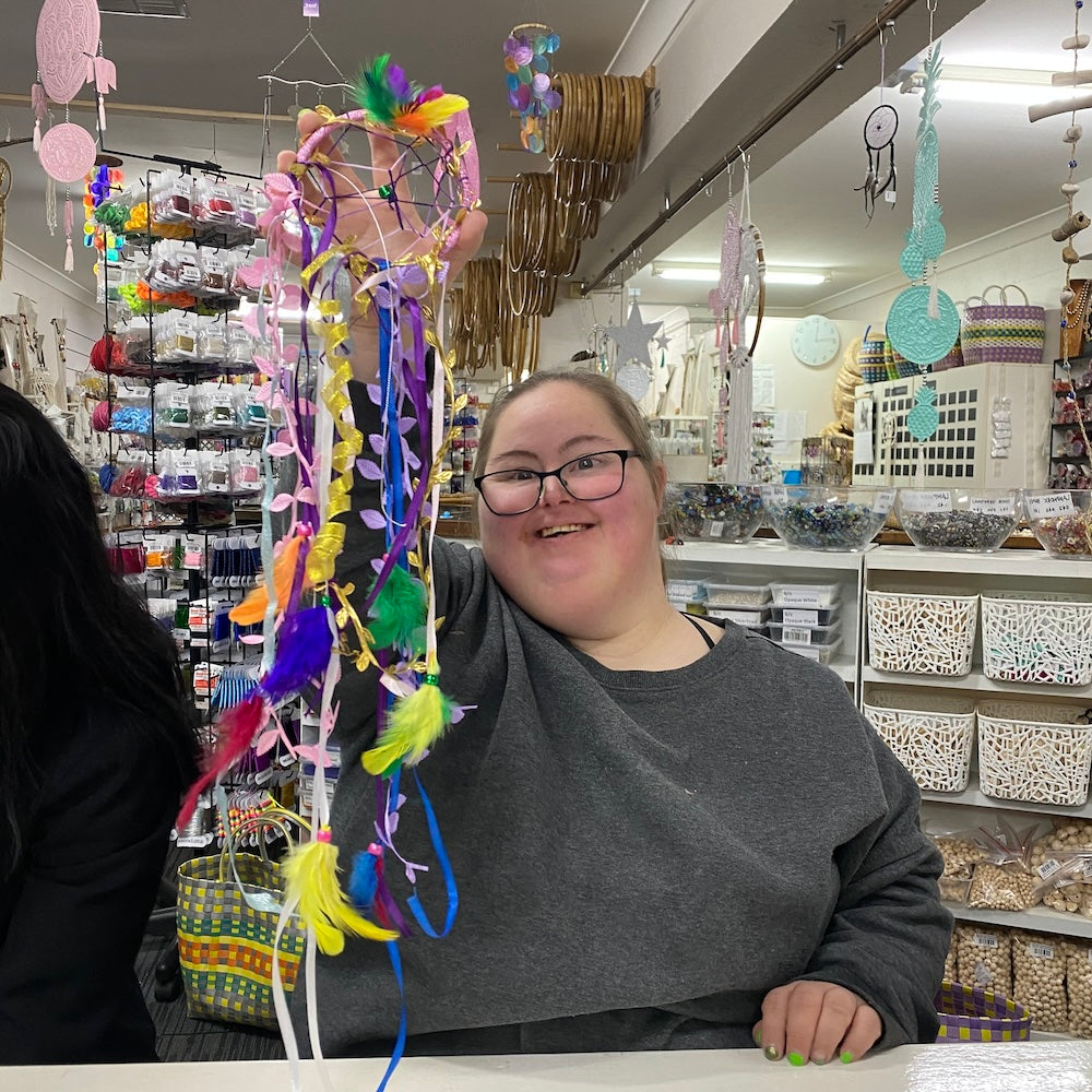An NDIS participant girl proudly displaying a colorful beaded craft creation at a workshop for disability showing a store background