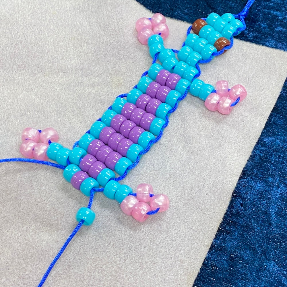 A beaded lizard workshop project not yet finished showing pink purple and blue beads with brown eyes on a cream and blue background