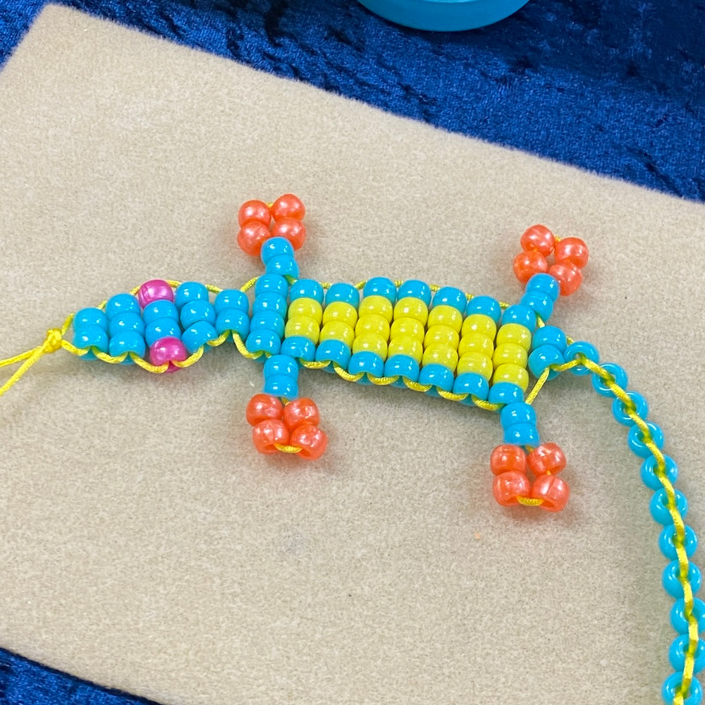 Gecko animal toy made from blue yellow and orange beads with pink eyes on a cream bead mat background