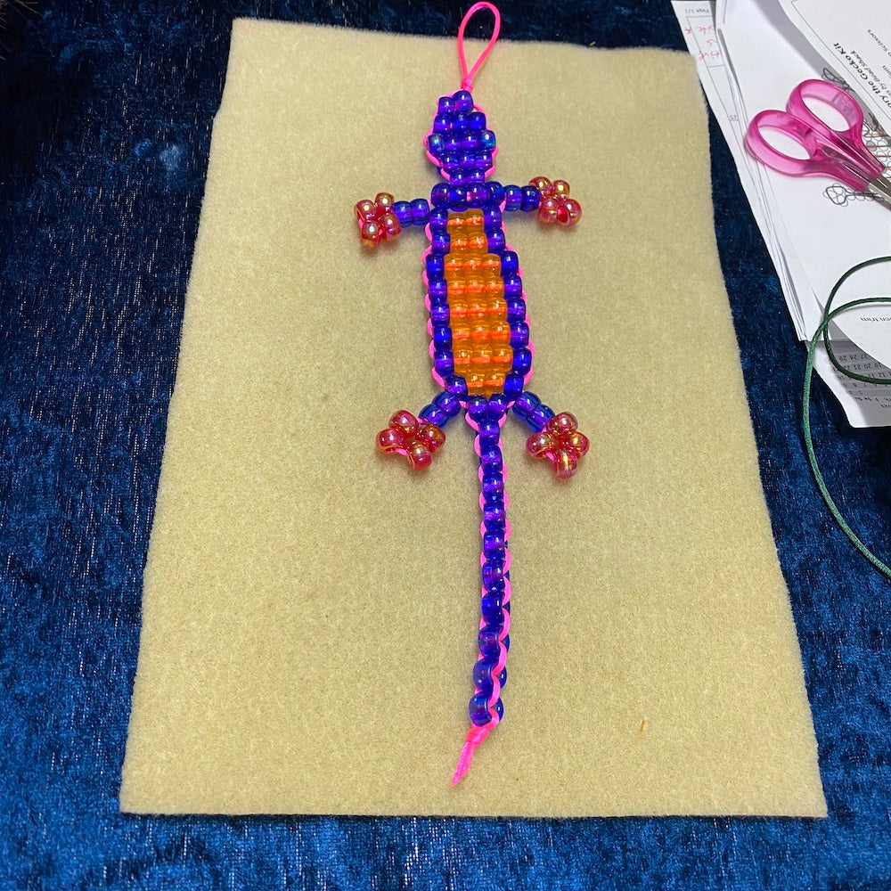 a lizard toy beaded from a kit in blue orange and red on a cream bead mat with scissors and paper in the background