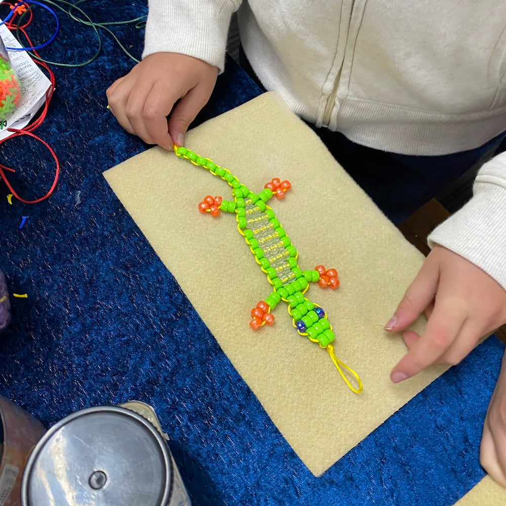 A child at a table showing a lizard beaded project using green yellow and orange beads with blue eyes on a cream bead mat on a blue tablecloth