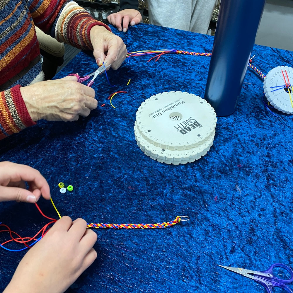 hands working on a Kumihimo weaving project key ring sat Bead Shack workshop table showing a blue background with a teacher using scissors to trim the cord