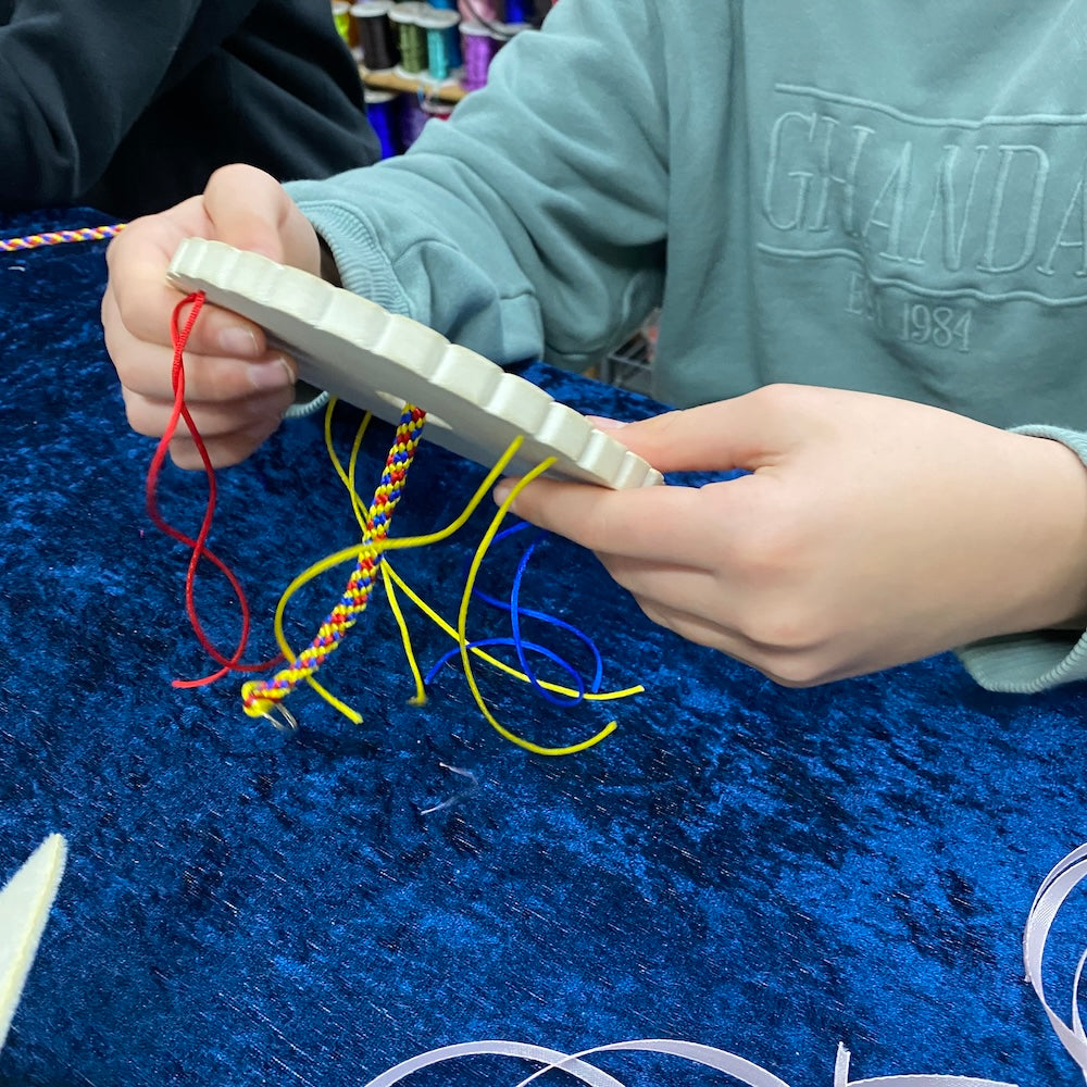 Child hands working on a Kumihimo weaving project key ring at Bead Shack workshop table showing a blue background