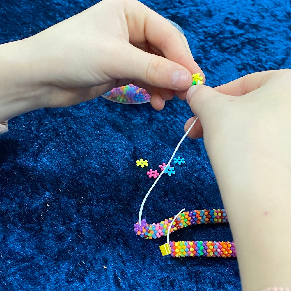 close up of a childs hands making a bracelet on white elastic string using colourful pinwheel beads in opaque colours on a blue background