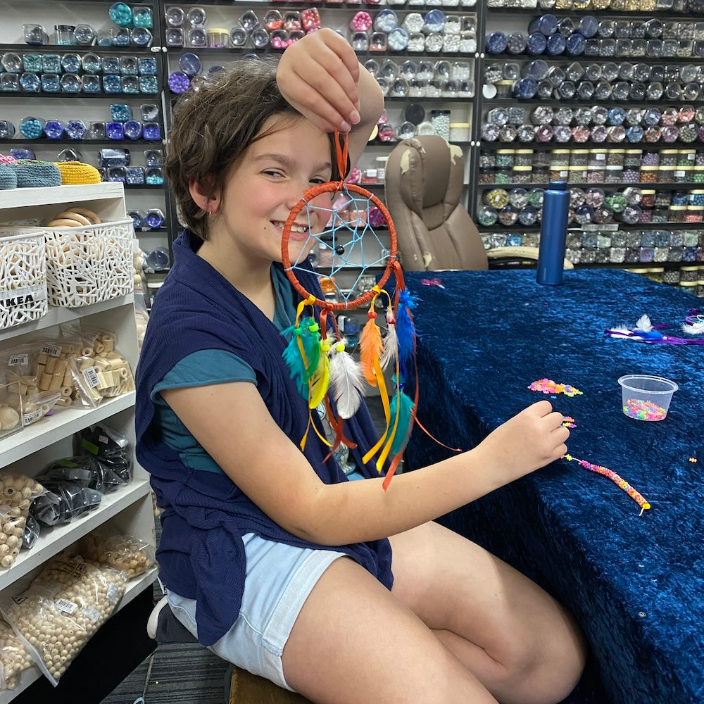 A child holding up a varied color dream catcher they have made, featuring bright feathers and beads with orange ribbon against a store background.