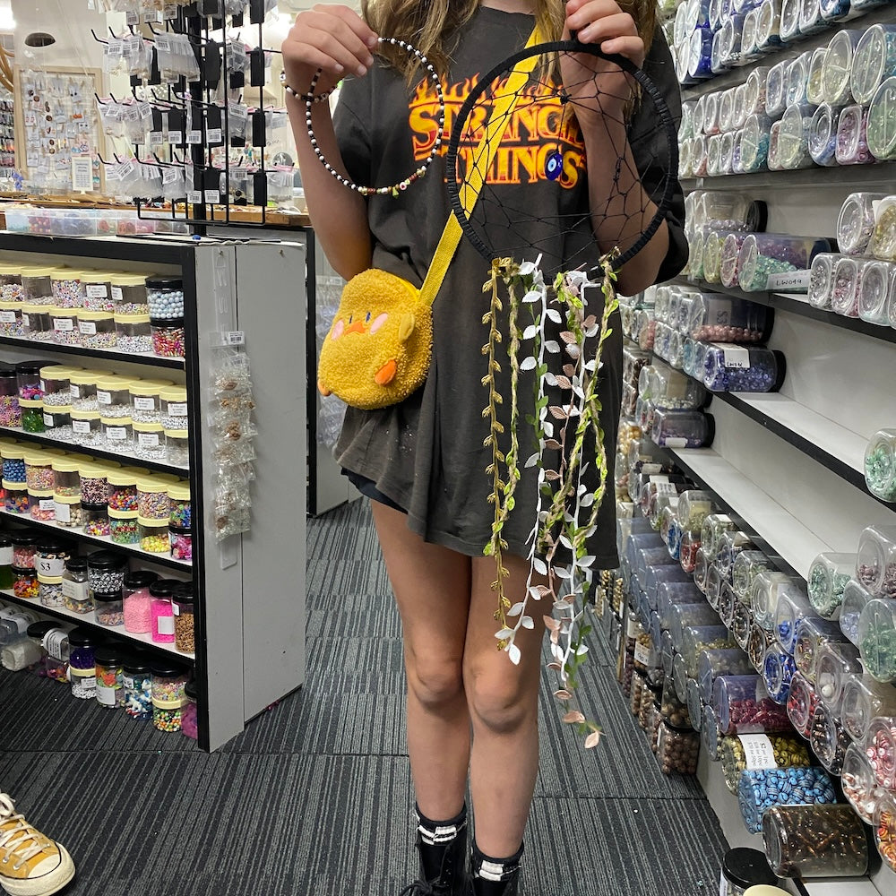 A child holding up a white dream catcher they have made, featuring black ribbon and web and evil eye centrepiece pendant beads and leaf braided cord against a store background.