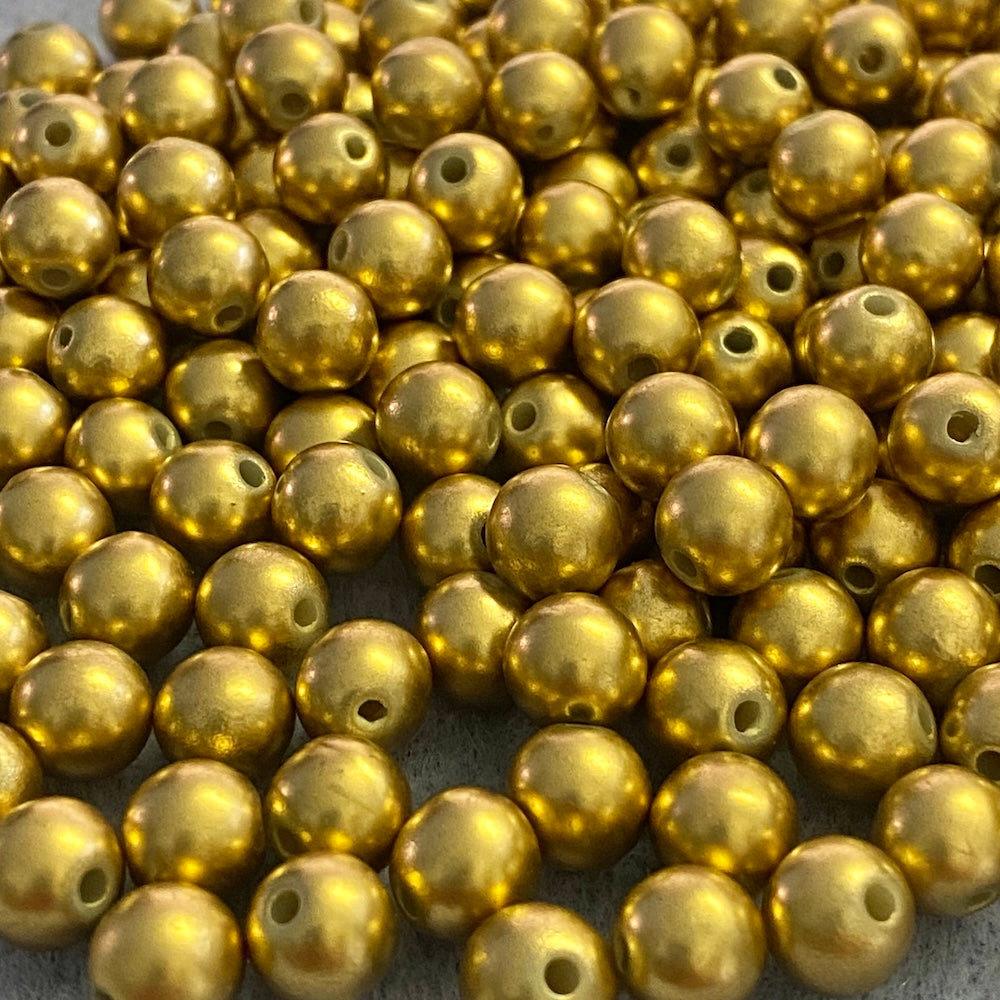A close-up pile of small metallic gold round beads with holes, filling the frame against a dark textured background.