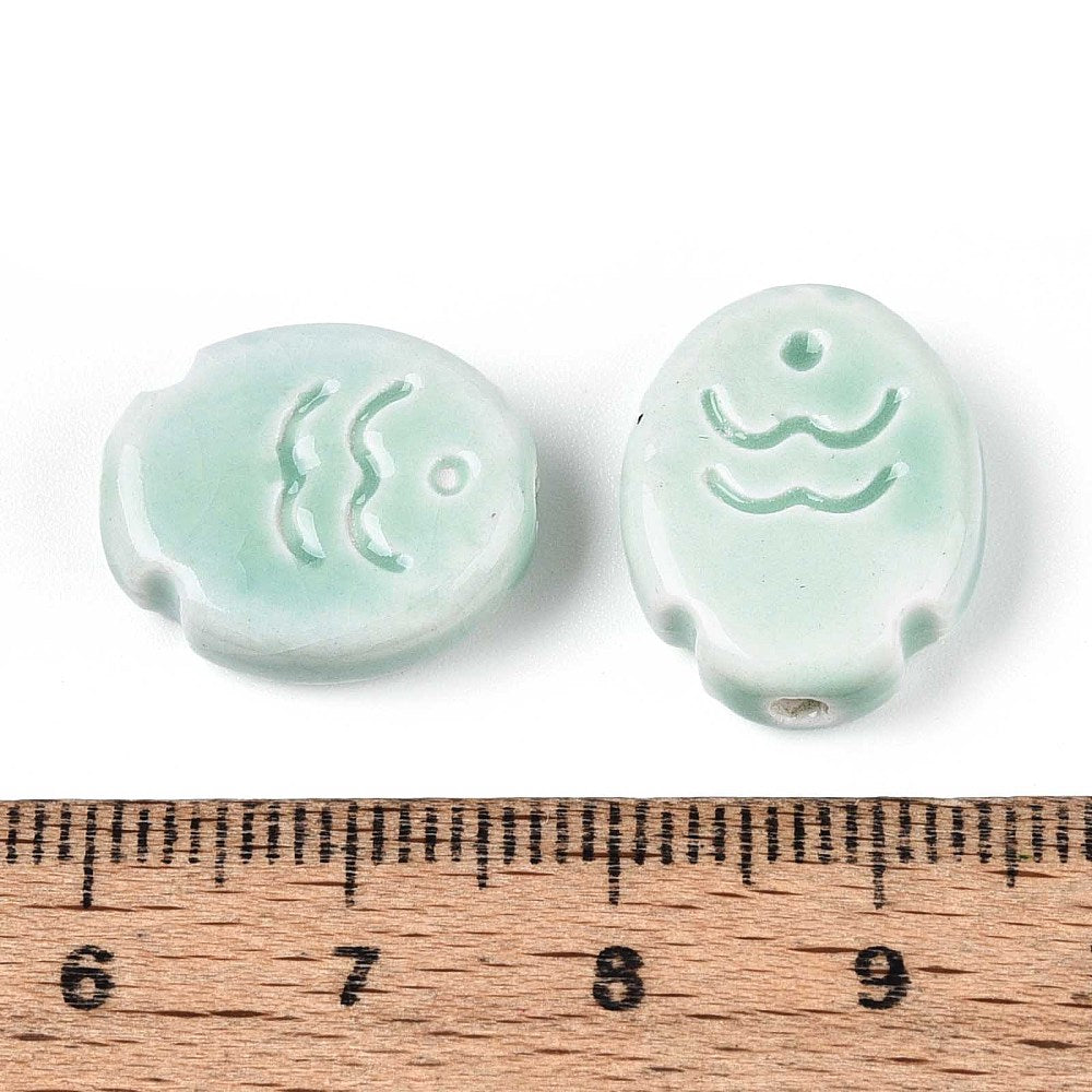Two light green ceramic fish beads with wave designs on a white background, measured against a wooden ruler.
