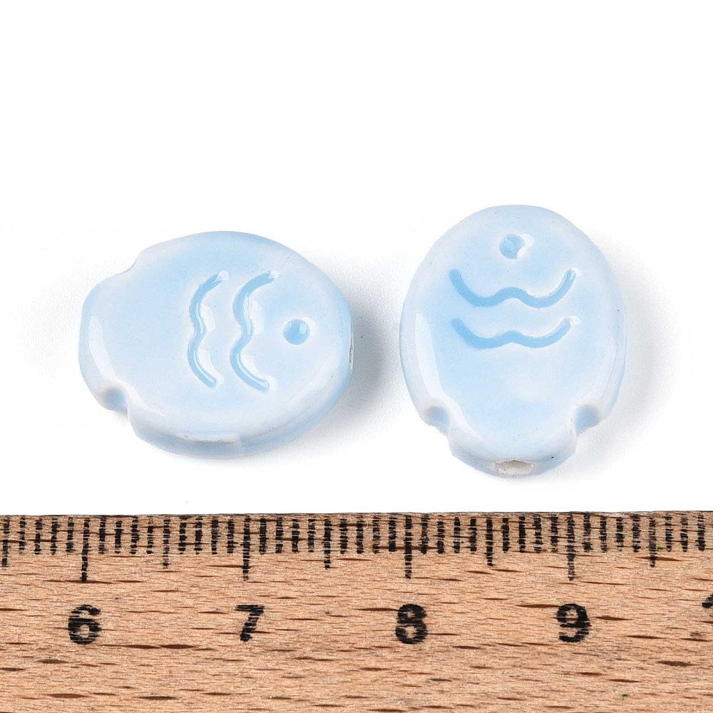 Two light blue fish ceramic beads with wave design on a wooden ruler for scale.