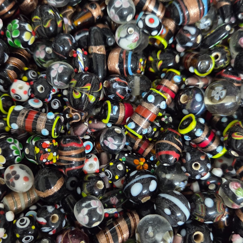 A dense pile of handmade glass lampwork beads featuring various patterns including stripes, polka dots, swirls, and floral motifs in black, brown, red, white, green, yellow, and blue.